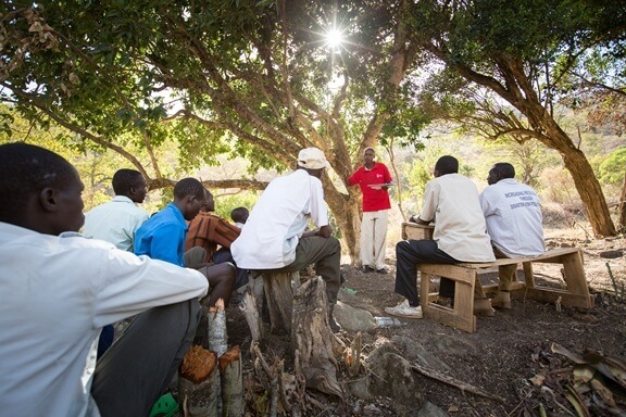 a group of men listen to a Red Cross worker teach under a tree