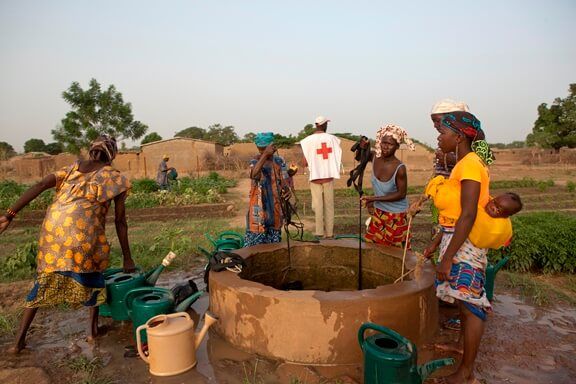 four women filling watering cans at a well while a Red Cross worker looks at field in background