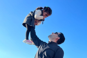 Amr holds his daughter Maria up in the air with one arm, with blue sky behind them.