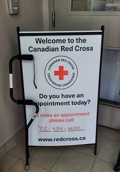 Inside the entrance of the Dartmouth, NS Red Cross office, a black offset cane rests vertically against a white Canadian Red Cross sign. The Red Cross logo is at the top and the black text reads: "Welcome to the Canadian Red Cross. Do you have an appointment today?"