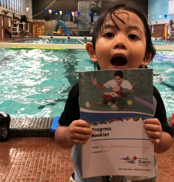 Kai smiling holding his Red Cross swim progress book infront of the pool