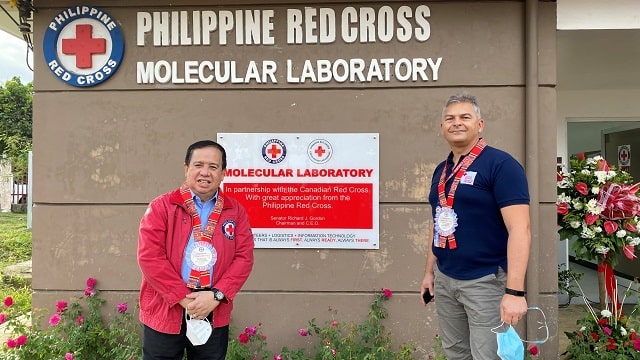 Two men standing in front of a large sign with text: Philippine Red Cross Molecular Laboratory