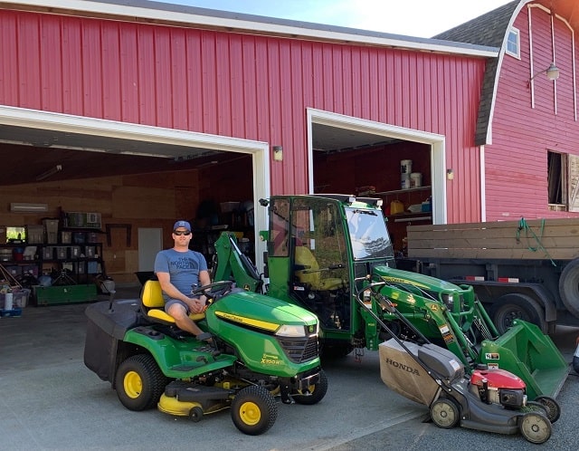 A man on a lawn mower with other landscaping vehicles in front of a large red barn