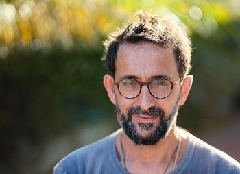 A man with black hair, a black and grey beard, and tanned skin is standing outdoors. He is wearing a blue shirt and glasses. He is smiling slightly.