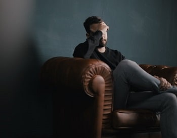 A man holding his hand up to his head sitting on a couch in darkened room. Photo credit: Nik Shuliahin/Unsplash