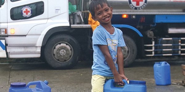 A small boy smiles at the camera as he carries a large blue water jug. a