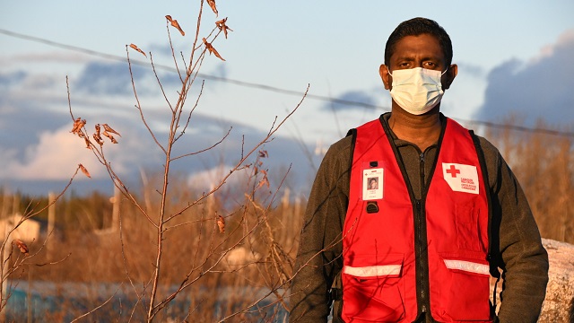 A Red Cross volunteer standing outdoors, wearing a surgical mask stares straight ahead.