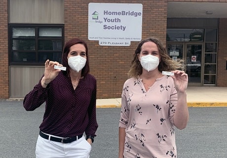Two women in masks holding rapid tests outside a building with a sign with text: HomeBridge Youth Society
