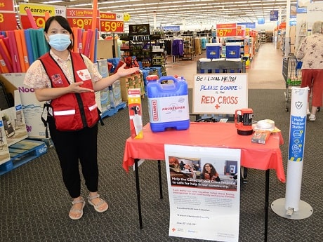 A woman in a Red Cross vest standing beside a display of emergency equipment such as batteries, flashlight and water container, facing the camera and smiling. The display is inside a Walmart store.