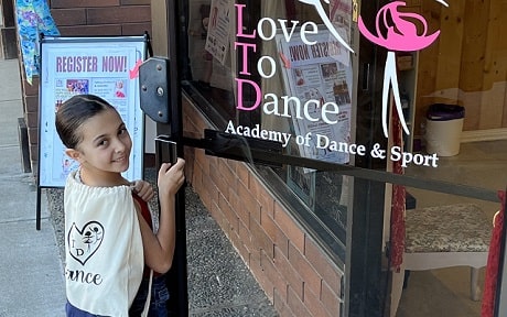 A young girl is opening the door to Love to Dance Academy studio while smiling at the camera