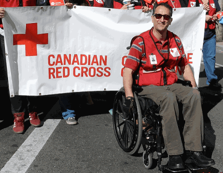 Mahmood in his wheelchair sitting in front of a Red Cross banner being held by other volunteers in Red Cross vests.