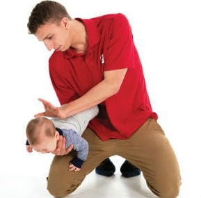 A man on his kness holds a baby downwards with his right hand, about to deliver a back blow with heel of his left hand