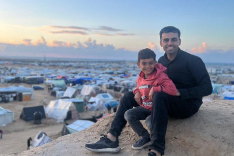 Amr and his son Adam sit on a hill, with tents in a camp for displaced people behind them
