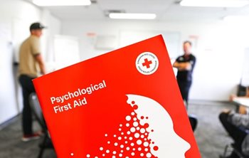 A close-up of a Psychological First Aid course booklet with people standing in the distance, blurred
