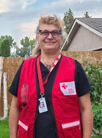 A woman stands in her backyard, wearing reading glasses and a bright red Red Cross vest, smiling.