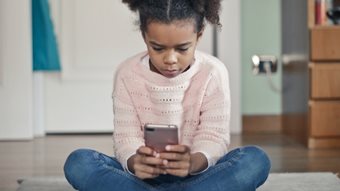 A young girl sitting on the floor of a bedroom, looking at a phone in her hands. Credit: bruce mars / unsplash.com