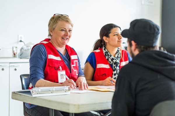 Two people wearing Red Cross vests speak to a person across a table. The workers are holding pens above paperwork on the table.
