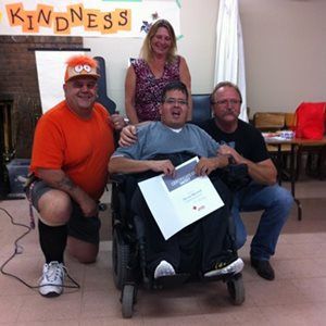 man in wheelchair with certificate surrounded by three people posing for camera