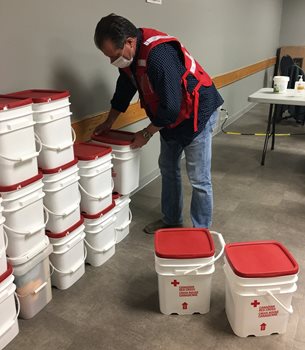 A man in mask and Red Cross vest picking up a Red Cross clean-up kit from a pile of many other kits lining a wall.
