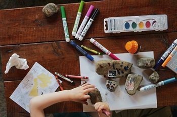 Paints and markers on a table with child's hands drawing on a rock
