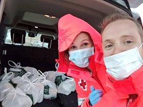 A man and a woman wearing medical masks pose with bags of groceries. The woman wears a red cross jacket.