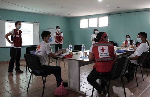 A group of people in Red Cross vests and masks sitting in a room on chairs in a circle