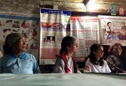 People in Nepal sitting and listening to someone speak. Posters about community health are hanging in the background.