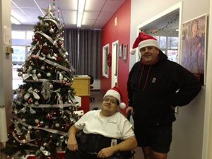 two men wearing Santa hats pose beside Christmas tree. One man is standing the other is in a wheelchair.