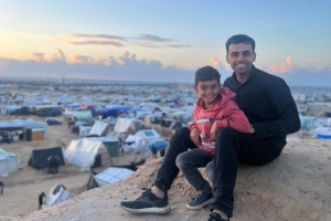 Amr and his son Adam sit on a hill, with tents in a camp for displaced people behind them.