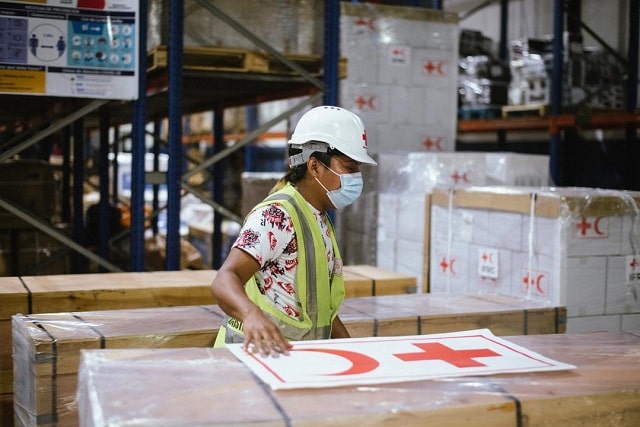 A person in helmet and mask standing among large boxes in a warehouse
