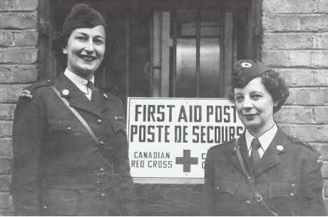 Two women in uniform from the 1940s standing in front of a Canadian Red Cross first aid post.