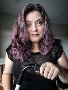 Jennifer Barnable, a woman with green eyes and purple-toned, wavy hair is standing in front of a white wall. She is wearing a black t-shirt and holding a black mobility aid in her left hand. She is facing the camera and is smiling slightly.