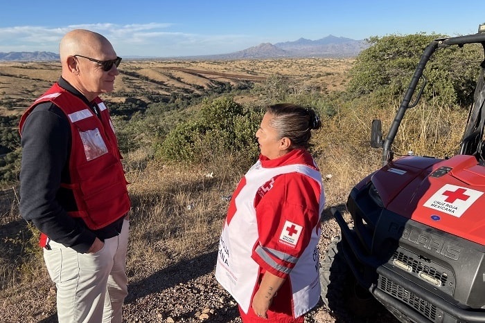 A man in a Red Cross vest and a woman in a Red Cross vest standing outside beside a small vehicle overlooking fields and hills in the background