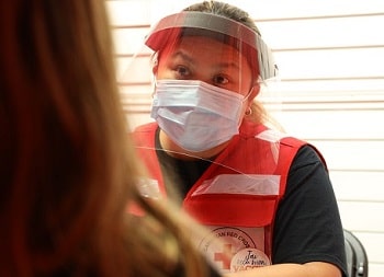 A woman in a mask and face shield, wearing a Red Cross vest,  speaking with someone