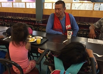 Tom Jackson in a Red Cross vest talking to two children sitting at a table.