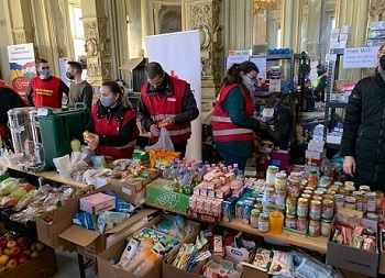 Masked Red Cross members behind tables full of food and supplies