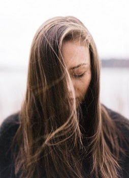 A woman with long, brown hair stands outside alone. She has her eyes closed.