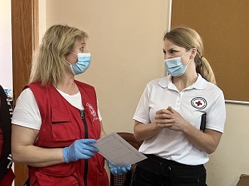 Two women in Red Cross shirts and masks stand together talking