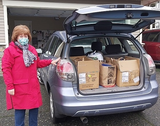 A woman in a mask standing beside a car full of boxes and supplies