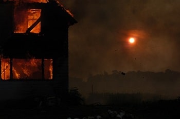 A burning house in a field at night with the moon shining through the smoke