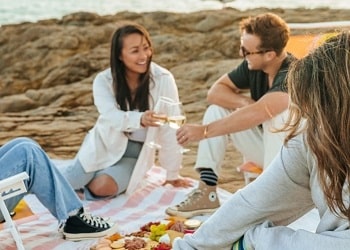 Two women and one man sit on a white blanket at the beach. They are laughing and talking while enjoying a picnic. A seagull sits in the background.