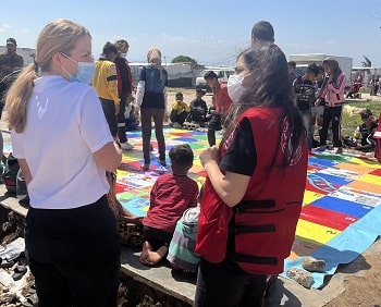 Two women in Red Cross shirts talking in the foreground while children play on a colourful mat in the background