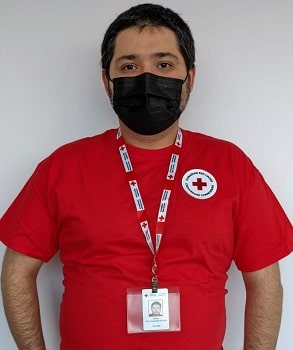 A  man in a mask, red shirt, and Red Cross lanyard with ID