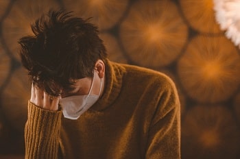 A man with dark hair and light skin is wearing a white covid mask and yellow sweater. He sits with his head in his hand, appearing emotional.