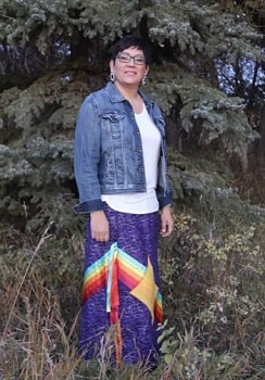 An Indigenous woman standing in front of large trees wearing a ribbon skirt and jean jacket.