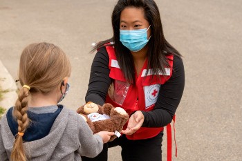 A woman in a mask and Red Cross vest offering a teddy bear to a young girl