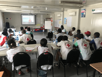 Several people sit around a table, all wearing white Red Cross shirts, watching a screen in front of the room.