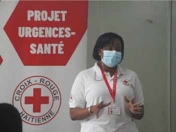 A woman wearing a mask and a white Red Cross sweater, standing in front of a sign with the Red Cross logo.