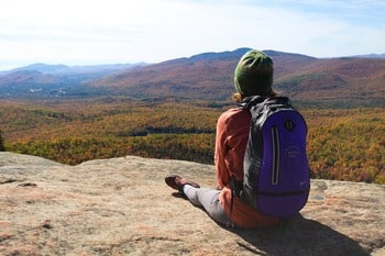 A woman with a green backpack, sitting on a rock, looking at the landscape of mountains with colorful trees