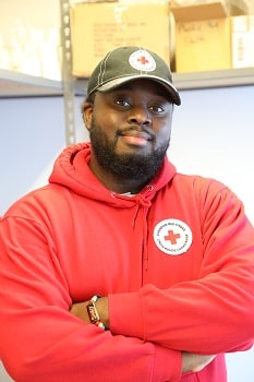 A man in Red Cross hat and sweater standing by shelves of supplies